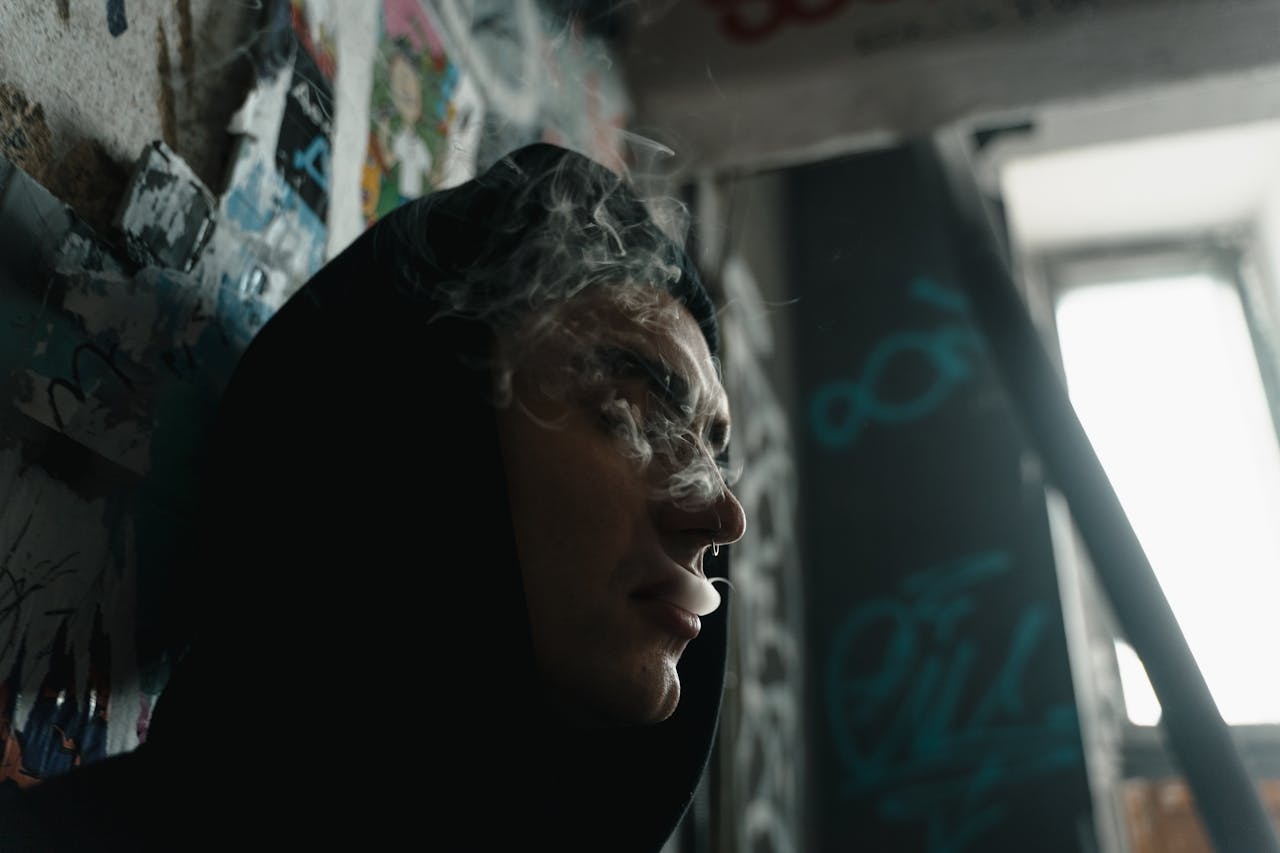 About Person in hoodie surrounded by graffiti, exhaling smoke in urban setting.